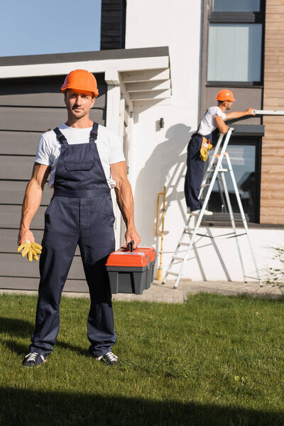 Selective focus of builder with gloves an toolbox standing on lawn while colleague working on ladder near house 