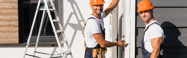 Panoramic concept of builders in helmets and uniform looking at camera while using spirit level on facade of house 