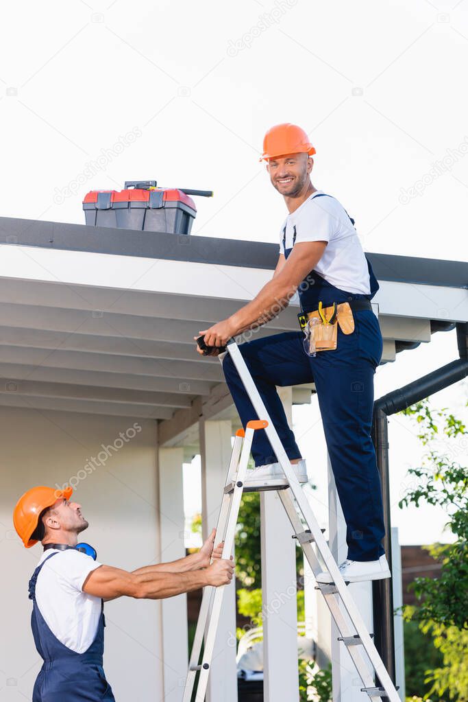 Side view of builder looking at camera while standing on ladder near colleague and building 