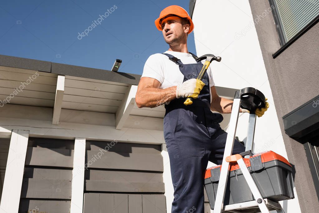 Builder in overalls and gloves holding hammer beside toolbox on ladder and building 