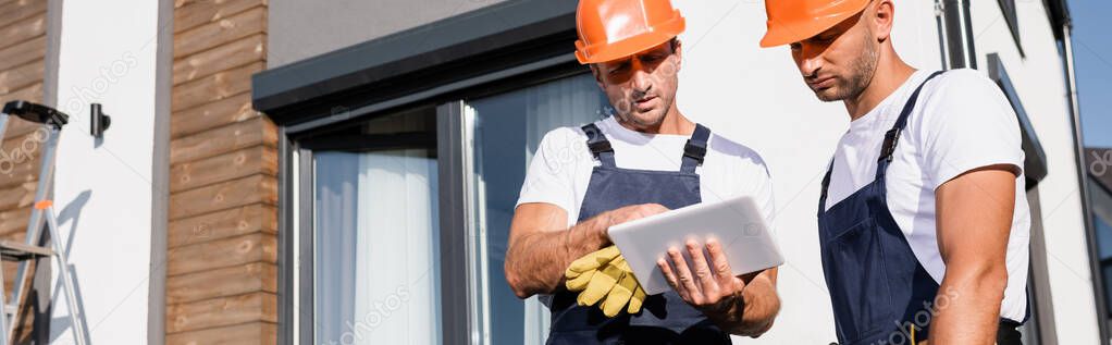 Horizontal image of workmen in hardhats and uniform using digital tablet near house 