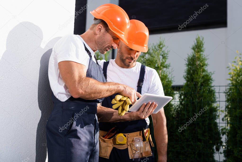 Handyman in overalls using digital tablet while standing near facade of house 
