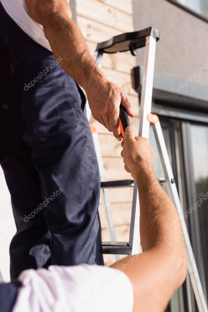 Cropped view of builder giving pliers to colleague on ladder near building 