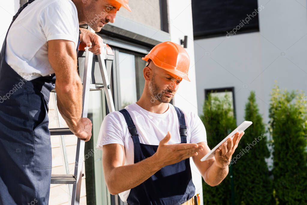 Selective focus of builder on ladder standing near colleague pointing with hand on digital tablet near building 