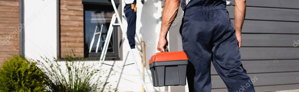 Panoramic crop of builder in uniform holding toolbox near house outdoors 