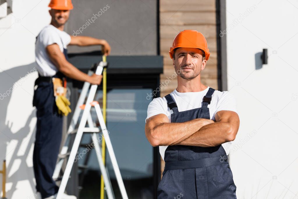 Selective focus of builder with crossed arms looking at camera with colleague and building at background 