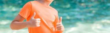 Kesilmiş turuncu tişörtlü çocuk başparmaklarını kaldırıyor, web sitesi başlığı