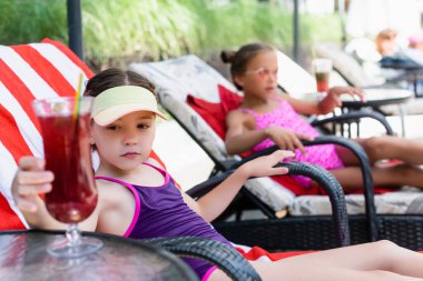 selective focus of girl in swimsuit and sun visor cap sitting in sunbed near friend and touching cocktail glass clipart
