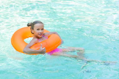 Şişme yüzükle havuzda yüzen ve baş parmağını gösteren mutlu bir kız.