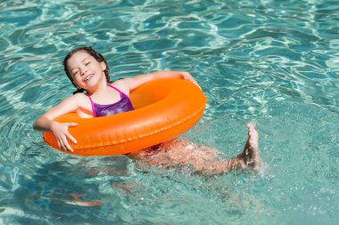 Şişme yüzükte havuzda yüzerken kameraya bakan mutlu kız.