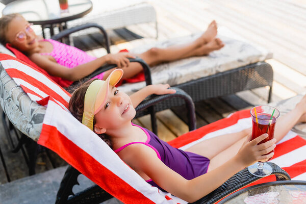 selective focus of girl in swimsuit holding glass of fresh cocktail while sitting in sunbeds near friend
