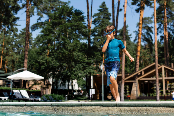 boy in t-shirt, shorts and swim googles plugging nose while standing at poolside and going to jump