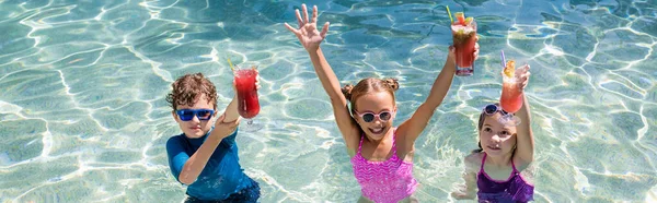 全景男孩和女孩站在游泳池里 举起双手举着清爽的鸡尾酒 — 图库照片