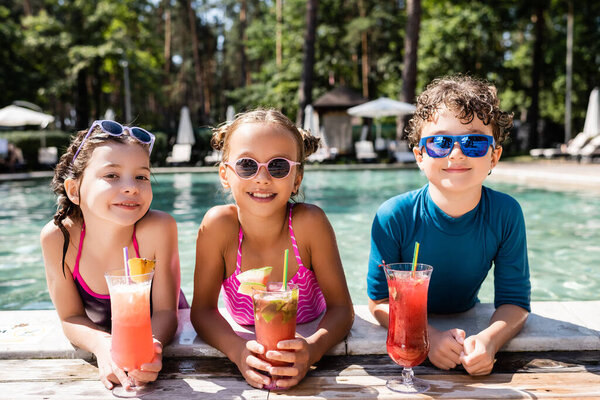 веселые друзья в купальниках смотрят в камеру, держа стаканы с фруктовыми коктейлями у бассейна