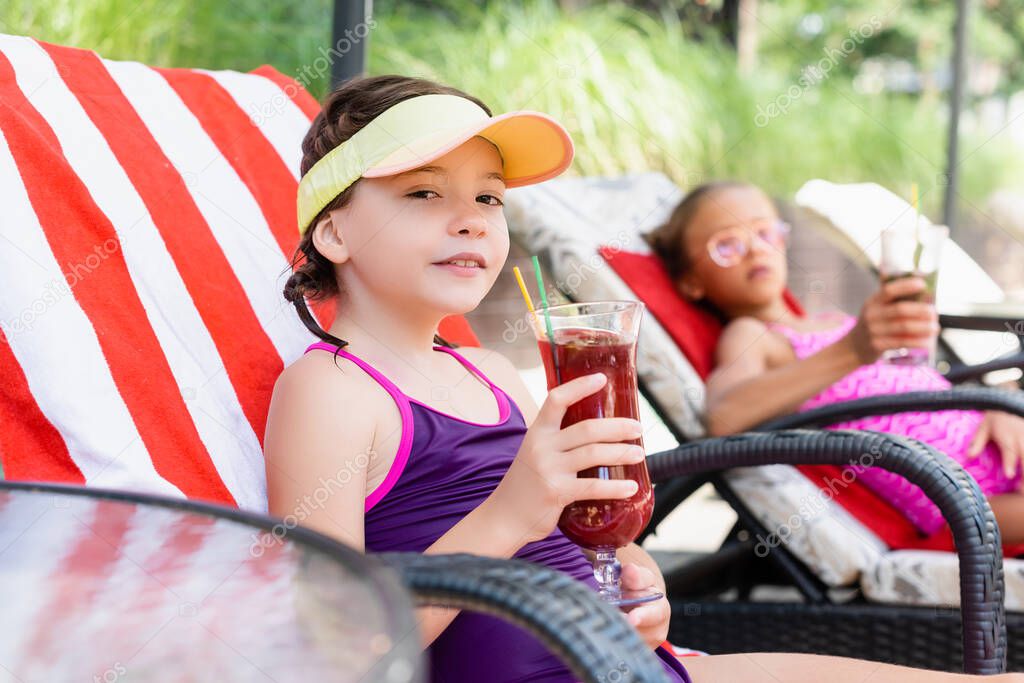 selective focus of girl in sun visor cap sitting in sunbed near friend and holding glass of fresh cocktail