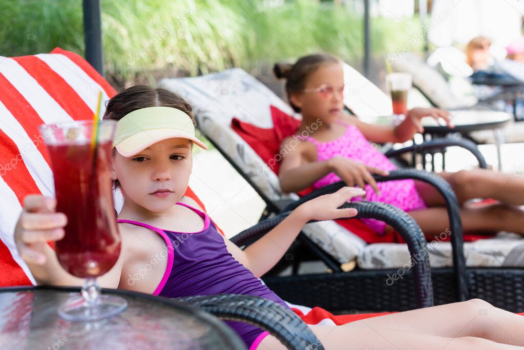 selective focus of girl in swimsuit and sun visor cap sitting in sunbed near friend and touching cocktail glass