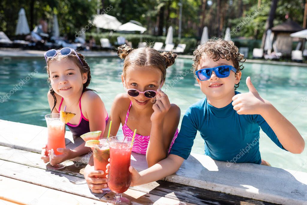 boy in sunglasses showing thumb up near girls with fresh fruit cocktails at poolside