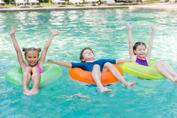 joyful girls with hands in air and boy with closed eyes floating in pool on swim rings