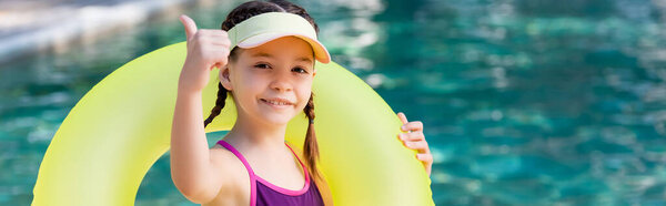 panoramic concept of girl in swimsuit and sun visor cap showing thumb up while holding swim ring 
