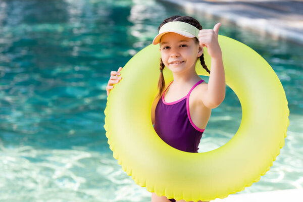 girl in swimwear and sun visor cap showing thumb up while holding swim ring near pool