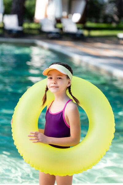 child in sun visor cap and swimsuit posing with inflatable ring near pool