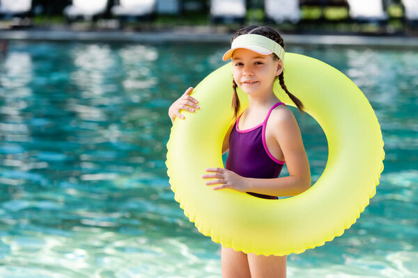 girl in swimsuit and sun visor cap holding inflatable ring while looking at camera near pool