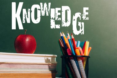 pen holder with color pencils, felt pens and ripe apple on books near green chalkboard with knowledge lettering  clipart