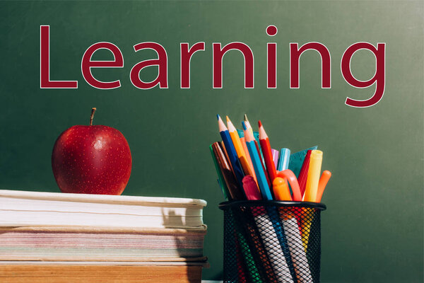 pen holder with color pencils, felt pens and ripe apple on books near green chalkboard with learning lettering 