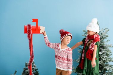 Şapkalı ve kazaklı bir çocuk, mavi üniformalı bir kız kardeşin yanındaki posta kutusuna zarf koyuyor.