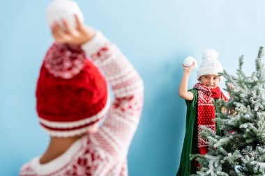 selective focus of kid in hat and scarf holding snowball while playing with brother on blue  clipart