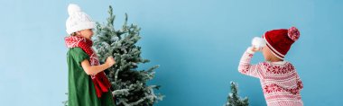 Kış kıyafetli çocukların mavi üzerinde Noel ağaçlarının yanında kartopu oynadığı panoramik bir fotoğraf.