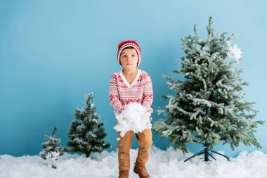 Kışlık kıyafetli çocuk, mavi Noel ağaçlarının yanında beyaz kar tutuyor.