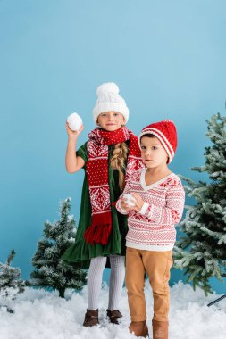 kids in winter outfit holding snowballs near christmas trees on blue clipart