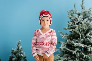 Kış kıyafetli çocuk mavi üzerinde Noel ağaçlarının yanında duruyor.