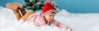 panoramik bir konsept. Kışlık kıyafetli bir çocuk. Beyaz kar üzerinde, mavi üzerinde Noel ağaçlarının yanında yatıyor.
