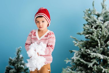 Kışlık kıyafetli çocuk, elinde soğuk karla, mavi Noel ağaçlarının yanında.
