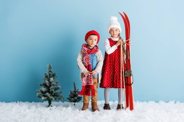Çocuğun elinde kayak sopası ve kayak takımı vardı. Kar üzerinde kardeşinin yanında duruyordu.