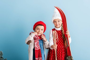 Kayak sopaları ve kayak takımları olan çocuk mavi şapkalı kardeşin yanında duruyordu.