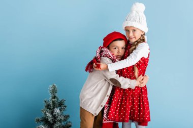 Kış kıyafetli çocuklar Noel ağacının yanında mavi renkte sarılıyorlar.