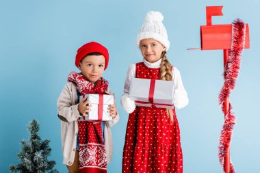 Çocuklar kışlık kıyafetleriyle posta kutusunun yanında hediyeler ve mavi üzerine köknar tutuyorlar.