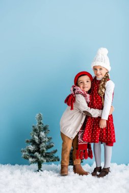 Kışlık kıyafetli çocuklar Noel ağacının yanında sarılıyorlar.