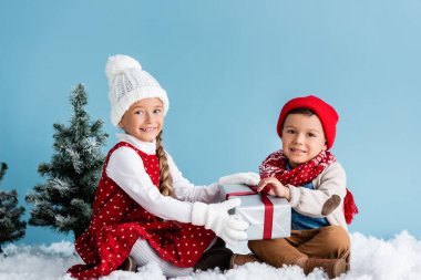Kışın giyinmiş çocuklar karda oturuyor ve mavi renkte izole edilmiş hediyelere dokunuyorlar.