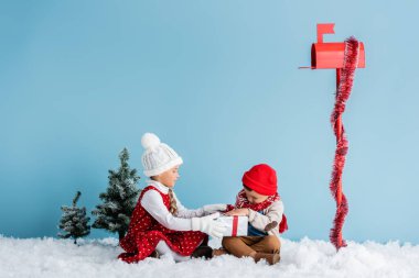 Çocuklar kışlık kıyafetleriyle karda oturuyor ve mavi posta kutusunun yanında hediyelere dokunuyorlar.