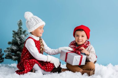 Şapkalı ve kışlıklı çocuklar karda oturuyor ve mavi üzerine izole edilmiş hediyelere dokunuyorlar.