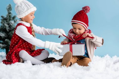 Şapkalı ve kışlıklı çocuklar karda oturuyor ve mavi üzerine izole edilmiş hediyelere dokunuyorlar.