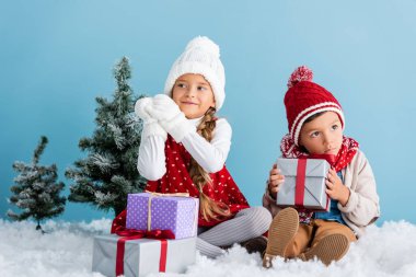 Kışın giysili çocuklar, karların üzerinde, köknar ve mavi renkli hediyelerin yanında oturuyorlar.