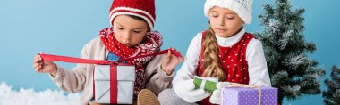 Kış kıyafetleri içinde çocukların karada oturduğu ve mavi renkte hediyeler tuttuğu panoramik bir çekim.