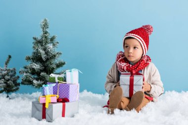 Şapkalı ve kışlıklı çocuk hediyelerin yanında karda oturuyor ve mavi üzerine izole edilmiş noel ağaçları.