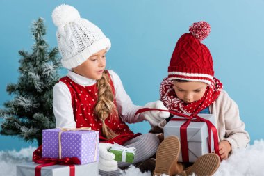 Kışlık kıyafetli çocuklar, karların üzerinde oturuyorlar ve mavi üzerine izole edilmiş hediyeler tutuyorlar.