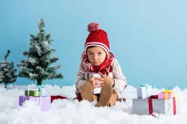 Şapkalı ve kışlıklı çocuk karda oturuyor ve mavi üzerine izole edilmiş Noel ağaçlarının yanında hediyeler tutuyor.